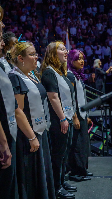 Members of the FSCJ Chorale singing the FSCJ Alma Mater while standing on stage, wearing black attire and white stoles monogramed with the FSCJ logo.