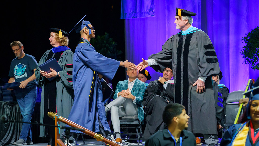 An FSCJ student wearing a navy-colored graduation gown is shaking hands with a member of the FSCJ District Board of Trustees who is wearing a black gown.