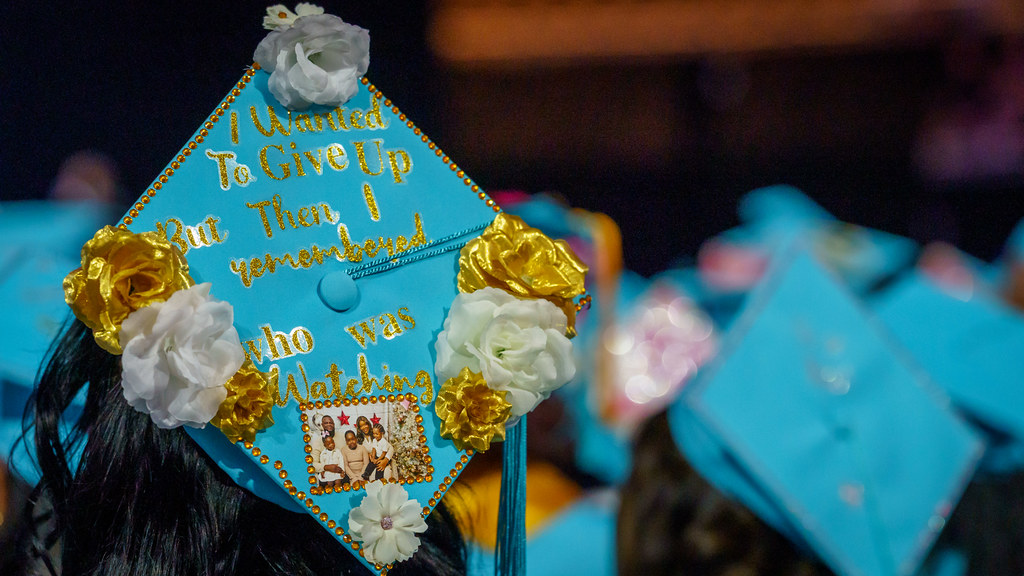 An FSCJ student is wearing a turquoise-colored cap decorated with gold-colored flowers, a family photo, and cursive text that reads