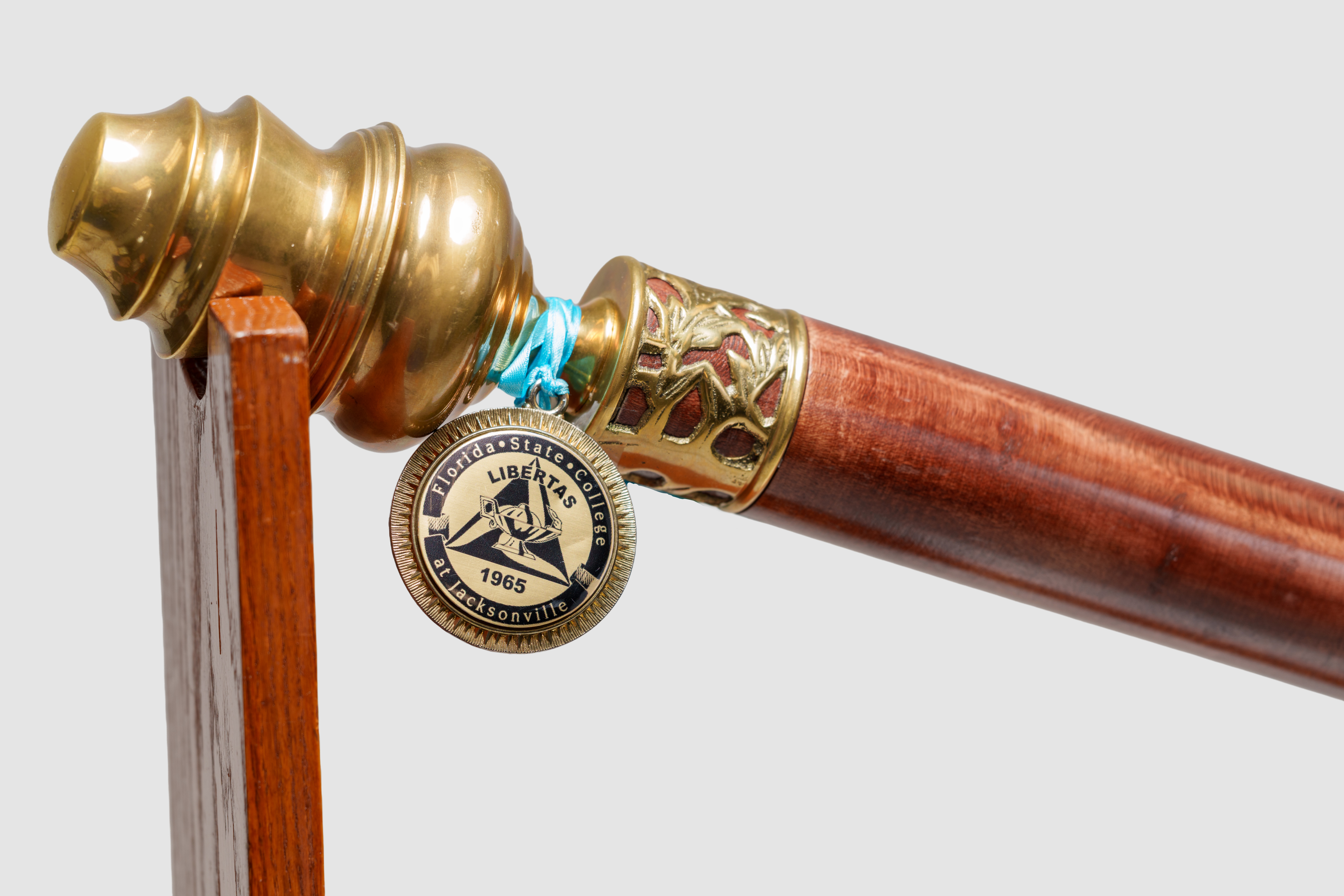 A close-up photo of the brass fitting header of the FSCJ Ceremonial Mace and a medallion with the FSCJ College Seal. The medallion is attached to the mace with a turquoise ribbon.