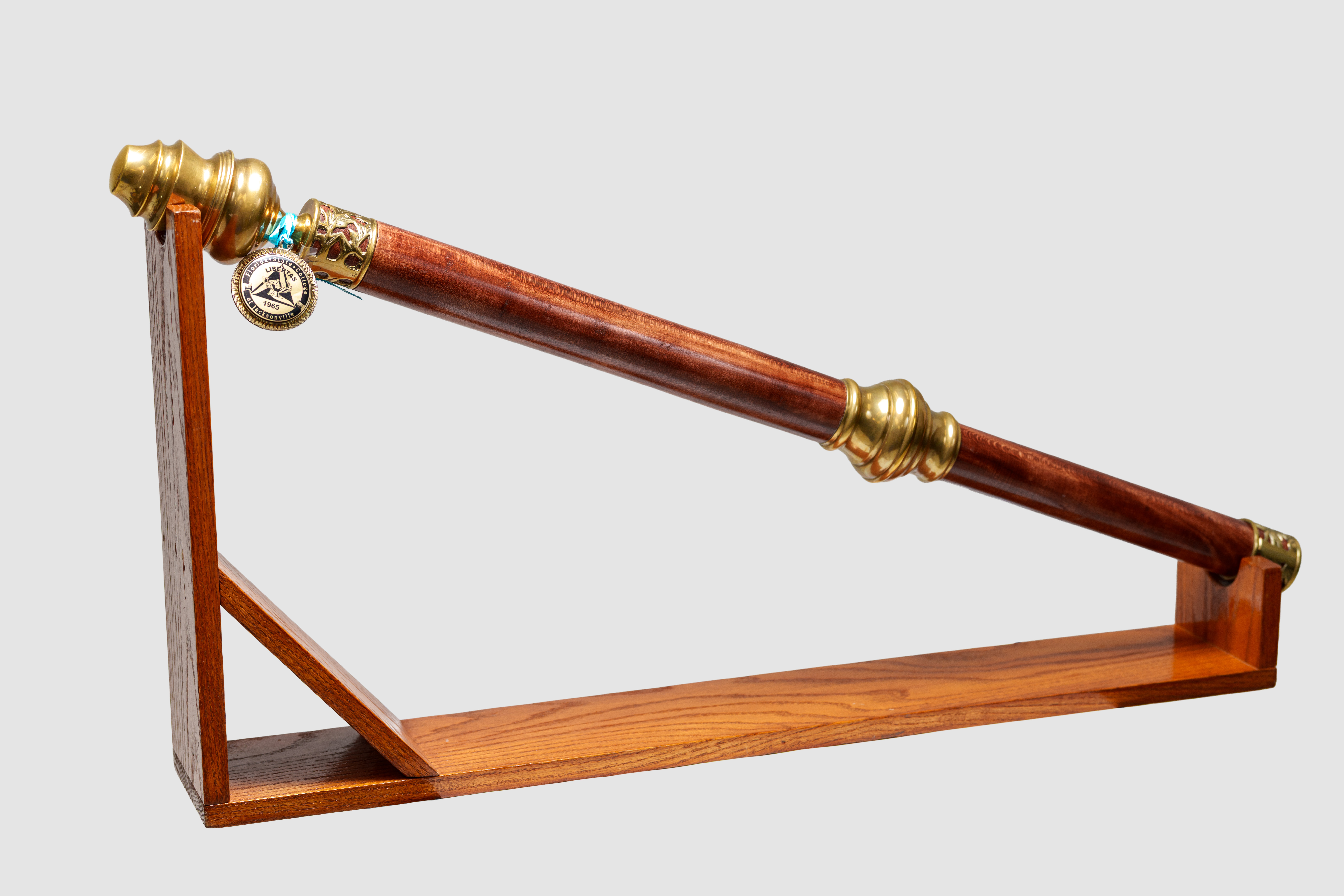 The FSCJ Ceremonial Mace is a wooden cylinder with brass fittings. A medallion with the FSCJ College Seal is attached with a turquoise ribbon. The mace rests on a wooden stand.
