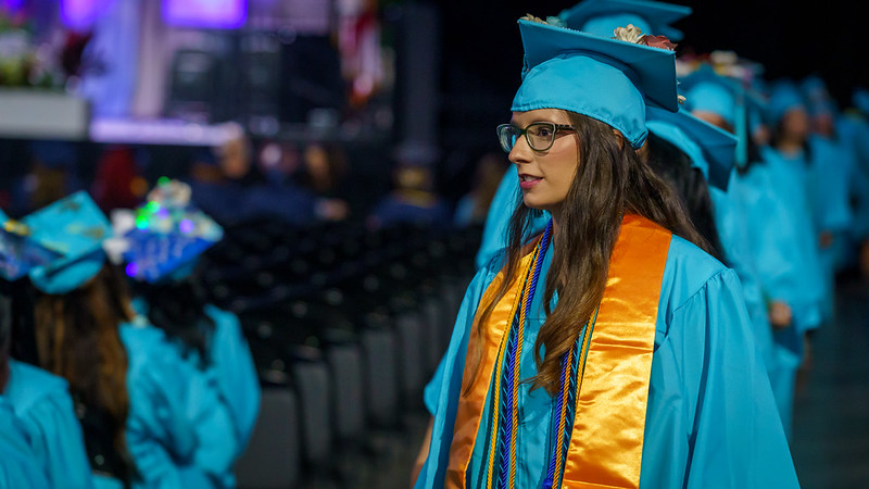 An FSCJ student is wearing a turquoise-colored cap and gown with a gold stole, gold cord, and a navy-blue and green-colored intertwined cord.