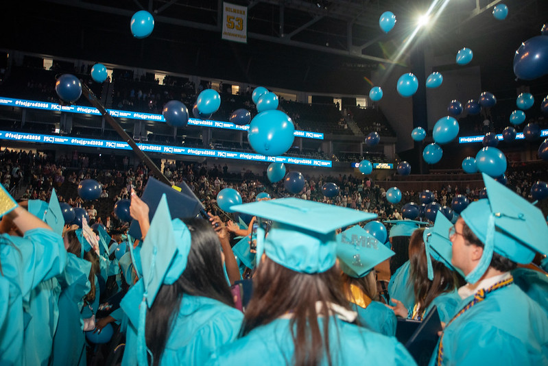 A group of FSCJ students in turquoise-colored caps and gowns throwing their caps in the air as turquoise and navy blue balloons fall from the ceiling.